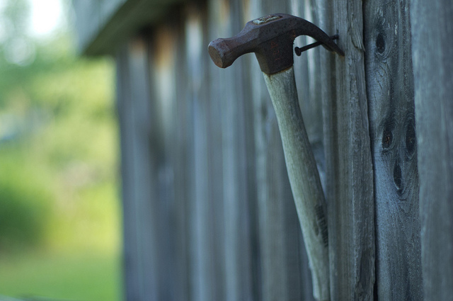 A hammer hangs on a nail sticking out of a wooden fence.